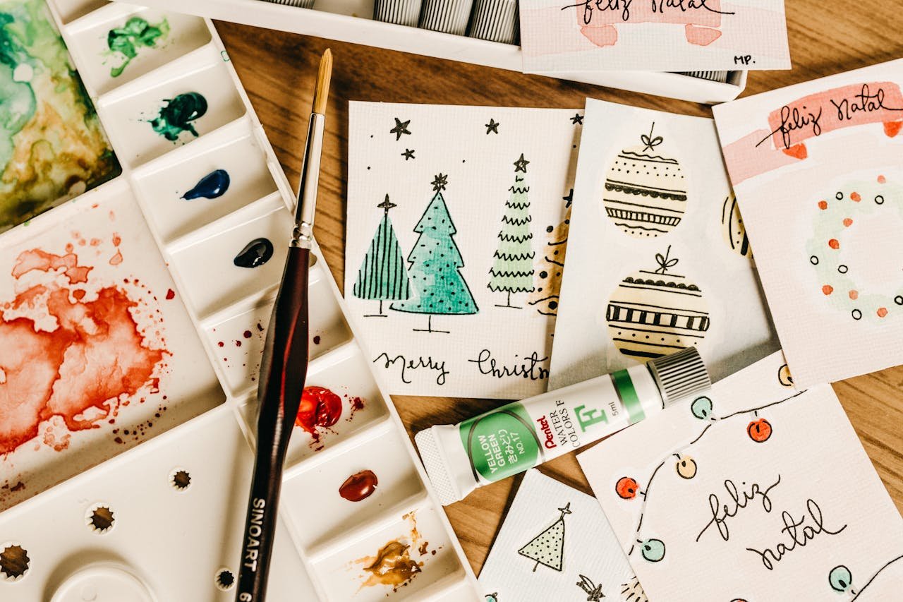 Home Artistic holiday card designs with watercolor paints and brushes on a wooden table.