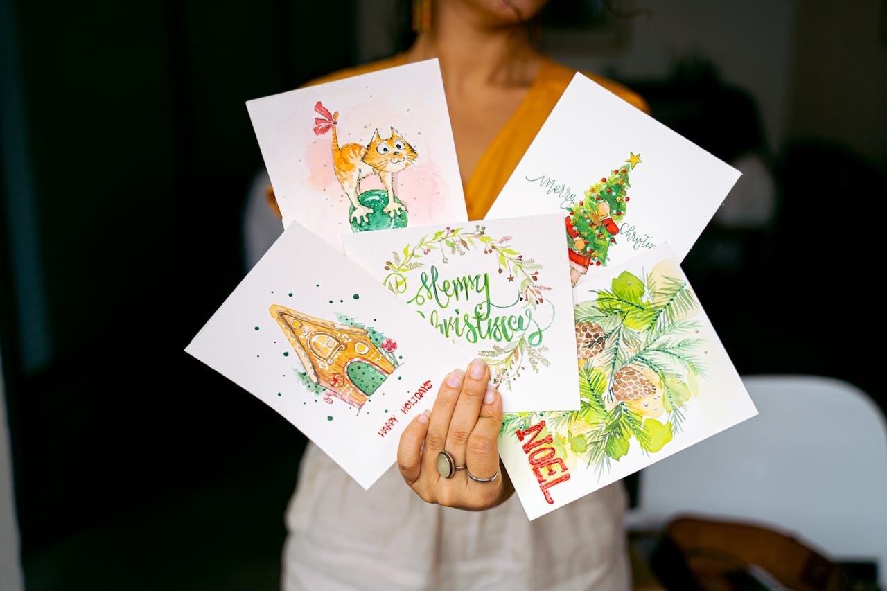 Home A collection of colorful Christmas cards held by a woman, featuring holiday-themed illustrations.